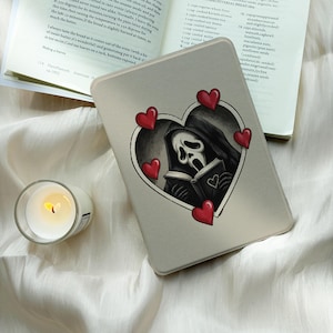 May include: A beige e-reader cover featuring a central heart-shaped frame containing a black and white illustration of a hooded figure with a mask reading a book, surrounded by five small red hearts, placed near an open cookbook and a candle.