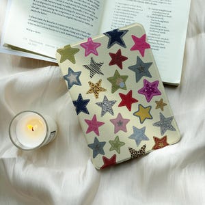 May include: A cream-colored e-reader case featuring a repeating pattern of stars in various colors and textures, including pink, red, blue, green, leopard print, and zebra print, positioned next to an open cookbook and a small glass candle.