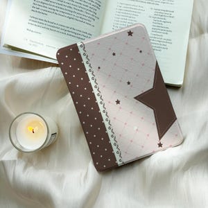 May include: A rectangular e-reader cover featuring a brown section with white stars and a light pink section with a grid pattern, small hearts, and stars. A decorative white lace-like border separates the two sections, and a large brown star is visible.