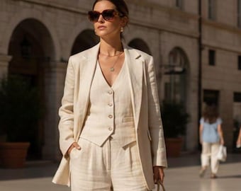 Pode incluir: Um fato de linho bege de três peças, composto por um blazer, colete e calças de perna larga. O fato é combinado com óculos de sol e uma pequena bolsa de mão tecida. O traje é usado ao ar livre em um ambiente urbano.