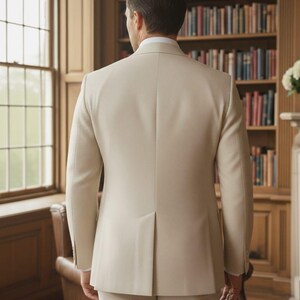 May include: A beige suit, consisting of a tailored jacket and matching trousers, is shown from the back. The jacket has a notched lapel and a vent in the back. The suit is worn over a white dress shirt. The background includes a window, a bookshelf, and a leather chair.