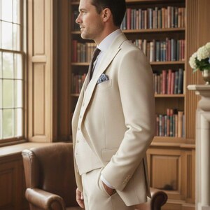 May include: A three-piece beige suit with a white dress shirt and patterned tie. The suit includes a jacket, vest, and trousers. A pocket square is visible in the jacket pocket. The background features a library with bookshelves and a leather armchair.