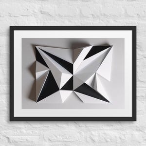 Könnte beinhalten: Gerahmter geometrischer abstrakter Kunstdruck. Das Kunstwerk zeigt ein 3D-Design mit schwarzen, weißen und grauen Dreiecksformen. Die gerahmte Kunst wird vor einer weißen Backsteinmauer ausgestellt.