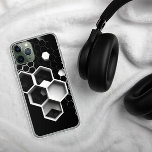Op de afbeelding: Een doorzichtige telefoonhoes met een zwart-wit honingraatontwerp. De hoes ligt naast een zwarte koptelefoon op een witte, getextureerde ondergrond. Het geometrische patroon is opvallend.