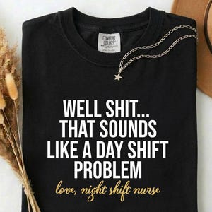 May include: Black t-shirt with the text "WELL SHIT... THAT SOUNDS LIKE A DAY SHIFT PROBLEM love, night shift nurse" in white and gold lettering. A silver necklace with a star pendant is on the shirt.