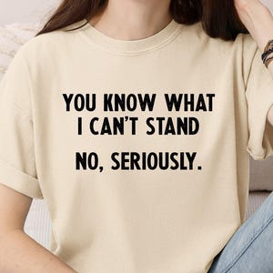 May include: Cream-colored t-shirt with the text "YOU KNOW WHAT I CAN'T STAND NO, SERIOUSLY." in black block letters. The shirt has rolled-up sleeves and is worn with blue jeans. The overall style is casual and relaxed.