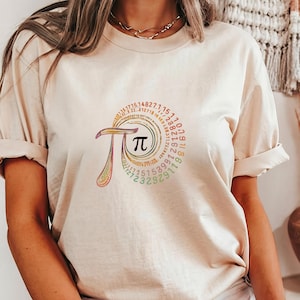 May include: Beige t-shirt featuring a colorful Pi symbol design. The design includes the Pi symbol in a circle, surrounded by a spiral of numbers. The symbol is in a variety of colors, including pink, green, and yellow.