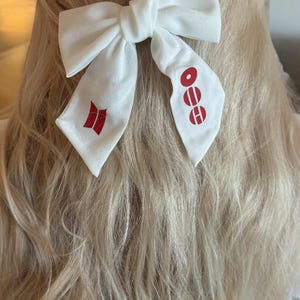 Può includere: Accessorio per capelli a forma di fiocco in tessuto bianco con dettagli grafici rossi. Il fiocco è annodato al centro e ha due lunghe code. Le code presentano disegni grafici rossi. L'accessorio per capelli è posto su uno sfondo di capelli biondi.