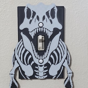 May include: A rectangular light switch cover plate featuring a light gray T-Rex skeleton silhouette layered over a solid black background. The design includes the skull, rib cage, and arms of the dinosaur, with a central toggle switch in white.