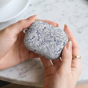 Könnte beinhalten: Eine Hülle für kabellose Ohrhörer mit weißem und blauem Blumenmuster. Die Hülle wird in zwei Händen gehalten, wobei ein goldenes Armband und ein Ring sichtbar sind. Eine Tasse Kaffee und eine Zeitschrift sind im Hintergrund.