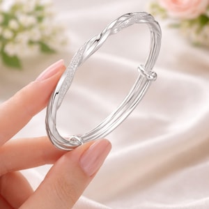 Peut inclure: Un bracelet jonc argenté avec un design torsadé. Le bracelet a une section texturée et scintillante et une finition lisse et polie. Le bracelet est réglable et comporte un fermoir simple. Le bijou est présenté sur un fond blanc doux.