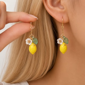 May include: A pair of gold-colored earrings featuring a yellow lemon charm with white flowers and green leaves. The earrings have a heart-shaped accent near the hook.