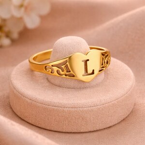 Op de afbeelding: Een goudkleurige ring met een hartvormig ontwerp met de letter "L" in het midden. De ring heeft decoratieve filigraanpatronen aan de zijkanten. De ring wordt gepresenteerd op een roze, cirkelvormige standaard.