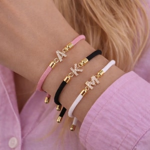 May include: Three initial bracelets are displayed, each with a different color cord: pink, black, and white. The gold-toned letters are encrusted with small, clear crystals. The bracelets are worn on a wrist, showcasing the personalized jewelry.