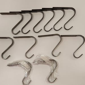 May include: A collection of dark metal S-shaped hooks, some individually wrapped in clear plastic. The hooks have a flat top section and a curved bottom, designed for hanging items. The hooks are likely for decorative or organizational purposes.