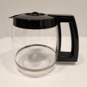 May include: A clear glass coffee carafe with a black plastic lid, handle, and spout. The carafe has measurement markings and a silver band around the base. The handle is rectangular.
