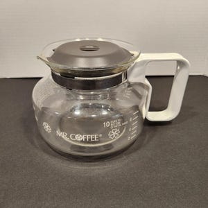 May include: Clear glass Mr. Coffee brand coffee carafe with a black lid and white handle. The carafe has a metal band and markings for up to 10 cups. The Mr. Coffee logo is visible on the front.