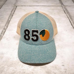 May include: A teal and tan trucker hat with the number "850" in black and an embroidered orange with green leaves. The hat has a mesh back and a curved brim.