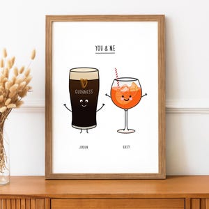 May include: A framed print featuring cartoon illustrations of a Guinness pint and an Aperol Spritz cocktail, each with arms and smiling faces. The text "YOU & ME" is above the drinks, with names "Jordan" and "Kirsty" below. The frame is brown.