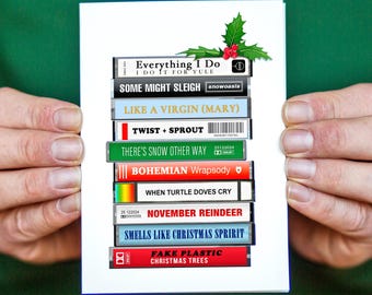 May include: A Christmas card with a stack of cassette tapes with song titles related to Christmas. The titles include "Everything I Do", "Some Might Sleigh", "Like a Virgin (Mary)", "Twist + Sprout", "There's Snow Other Way", "Bohemian Wrapsody", "When Turtle Doves Cry", "November Reindeer", "Smells Like Christmas Spirit", and "Fake Plastic Christmas Trees".