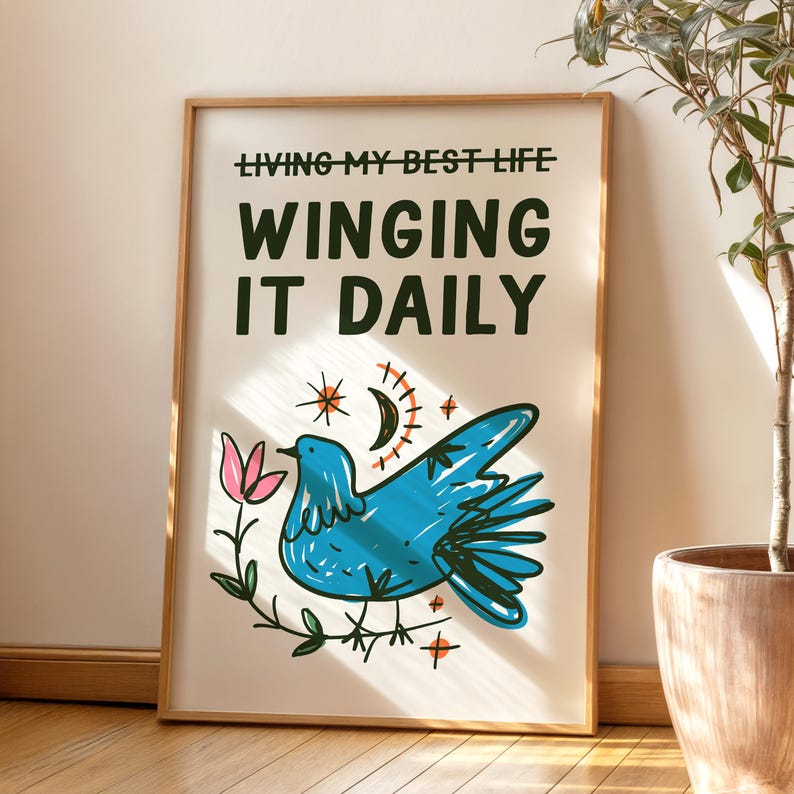 May include: A framed art print with the text "LIVING MY BEST LIFE WINGING IT DAILY." The artwork features a blue bird, a pink flower, and celestial elements. The frame is a light brown color.