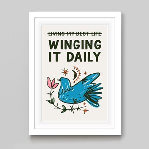 May include: A framed art print with the text "LIVING MY BEST LIFE WINGING IT DAILY." The artwork features a blue bird, a pink flower, a crescent moon, and stars. The art is in a white frame.