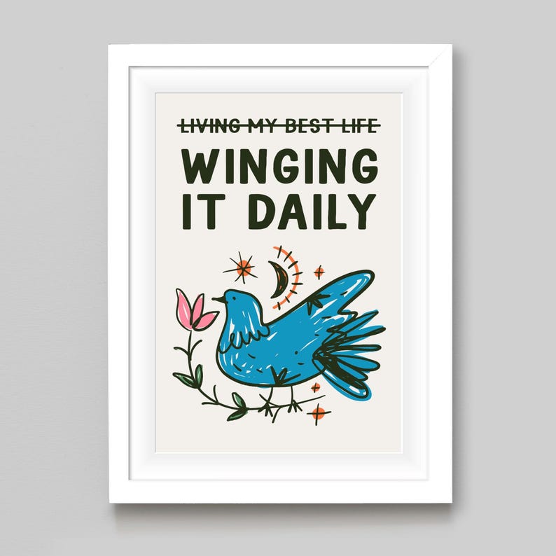 May include: A framed art print with the text "LIVING MY BEST LIFE WINGING IT DAILY." The artwork features a blue bird, a pink flower, a crescent moon, and stars. The art is in a white frame.