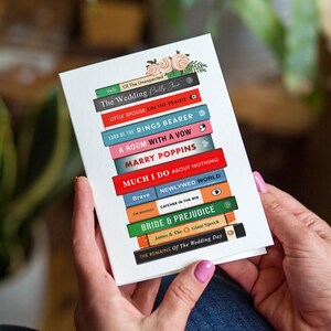 Pode incluir: Um cart&atilde;o branco com uma pilha de livros coloridos, incluindo "The Wedding Bells Jar" e "Bride & Prejudice". Detalhes florais no topo. O cart&atilde;o &eacute; segurado.