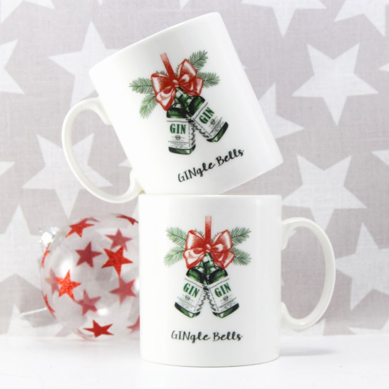May include: Two white ceramic mugs with a festive design. Each mug displays a pair of green gin bottles adorned with a red bow and pine sprigs, with the text "GIN" and "Gingle Bells". A clear bauble with red stars is in the foreground.