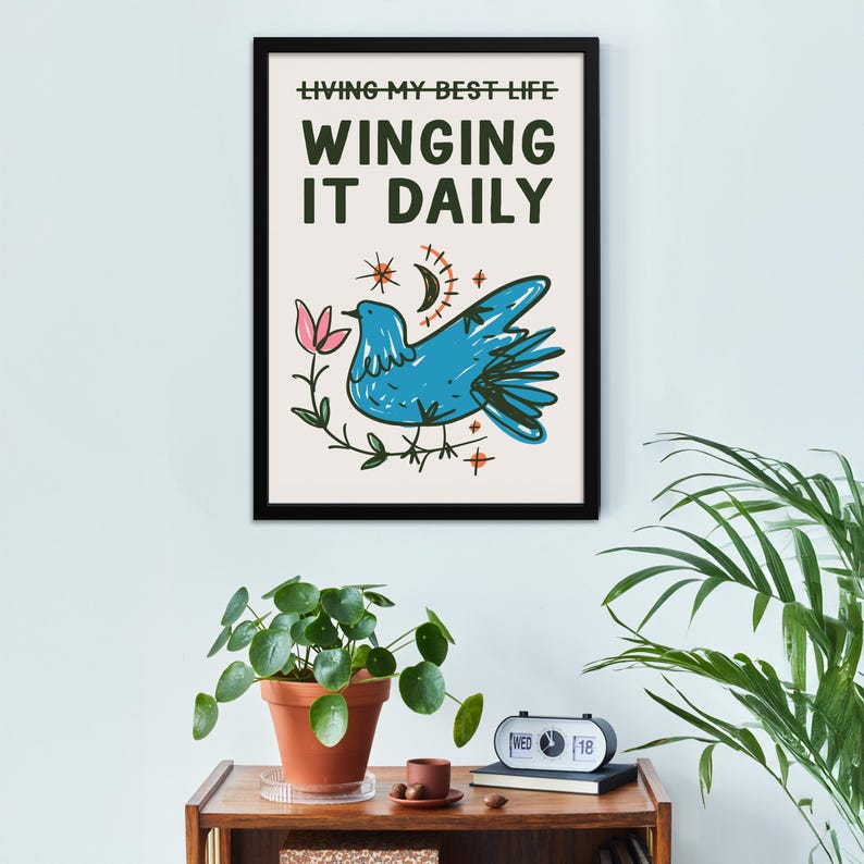 May include: A framed art print with the text "LIVING MY BEST LIFE WINGING IT DAILY." The artwork features a blue bird, pink flower, crescent moon, and stars. The print is in a black frame and hangs on a white wall.