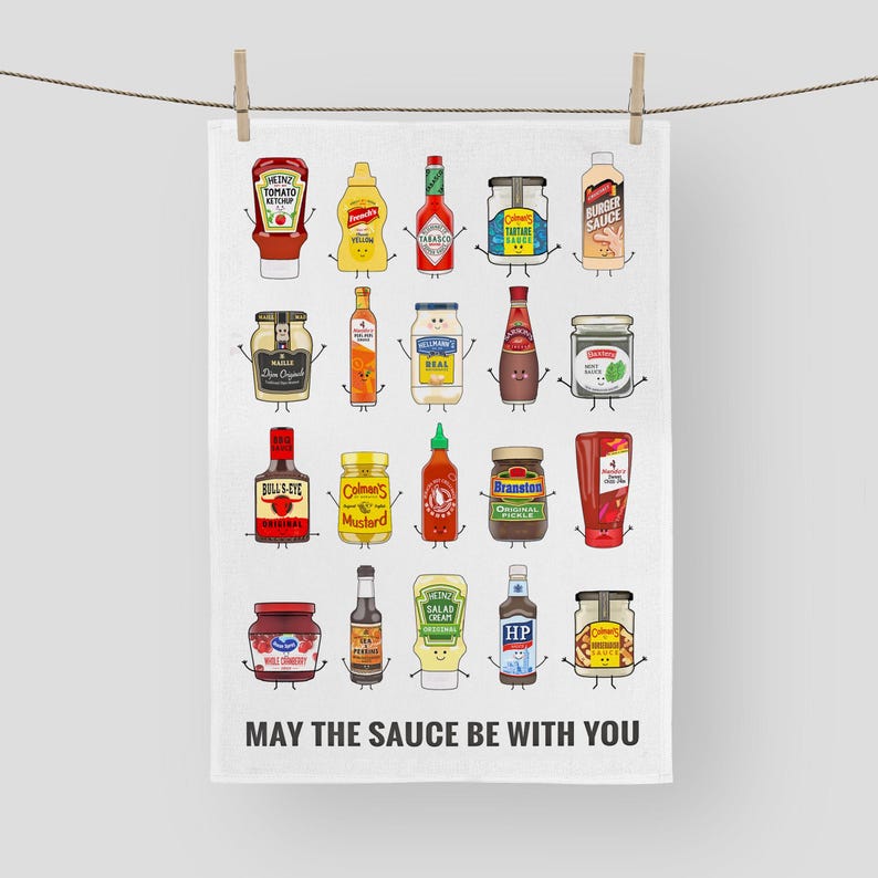 Peut inclure: Une serviette de cuisine blanche avec une illustration color&eacute;e de diverses bouteilles et bocaux de condiments. Les bouteilles et les bocaux sont dispos&eacute;s en rang&eacute;es et ont des visages de dessin anim&eacute;. Le texte "MAY THE SAUCE BE WITH YOU" est imprim&eacute; en bas de la serviette.