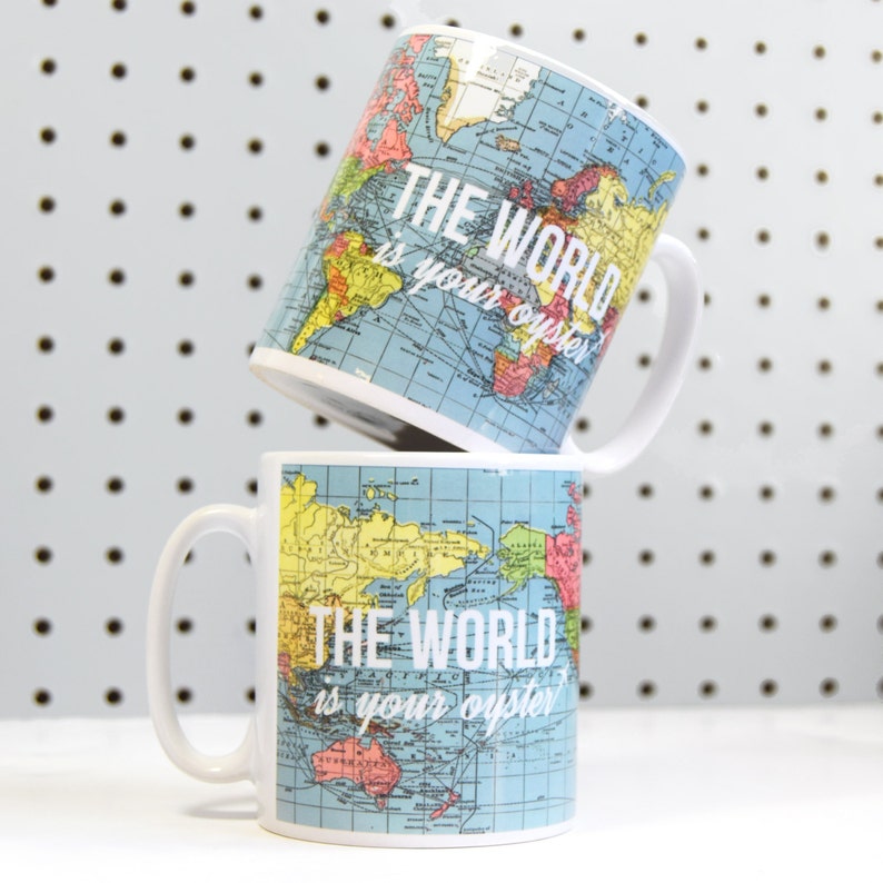 May include: Two white mugs with a world map design and the text "The World is your oyster" printed on them. The mugs are stacked on top of each other.