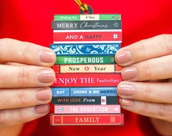 May include: A Christmas ornament shaped like a stack of books, held against a red background. The books have festive messages like "Merry Christmas" and "Enjoy the Festivities." A gold string is attached for hanging. The ornament is a unique holiday decoration.