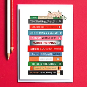 Pode incluir: Um cart&atilde;o branco com uma pilha de t&iacute;tulos de livros coloridos, incluindo "Bride & Prejudice" e "Marry Poppins". O cart&atilde;o &eacute; decorado com detalhes florais e l&aacute;pis sobre um fundo vermelho. Os t&iacute;tulos s&atilde;o empilhados verticalmente.