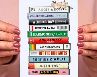 May include: A white card featuring a stack of cassette tapes with various text, including "Angie & Ross," "Wedding Day 28/08/25," and "With Love." Floral accents adorn the top. The card is held by hands with pink nail polish.