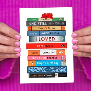 May include: A white greeting card featuring a stack of colorful book spines with text like "Happy Birthday" and "Absolute Classic." A red bow sits atop the stack. The card is held by hands with pink nail polish.