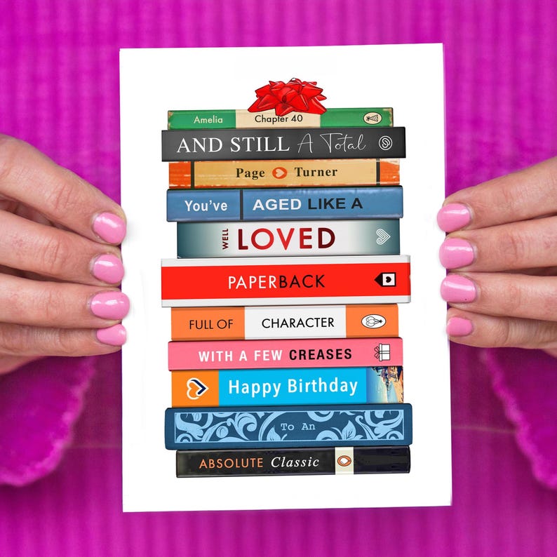May include: A white greeting card featuring a stack of colorful book spines with text like "Happy Birthday" and "Absolute Classic." A red bow sits atop the stack. The card is held by hands with pink nail polish.