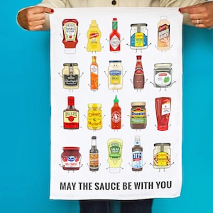 Peut inclure: Une serviette de cuisine blanche avec une illustration amusante de diverses bouteilles et bocaux de condiments avec des visages. Le texte "MAY THE SAUCE BE WITH YOU" est imprim&eacute; en bas.