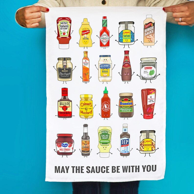 Peut inclure: Une serviette de cuisine blanche avec une illustration amusante de diverses bouteilles et bocaux de condiments avec des visages. Le texte "MAY THE SAUCE BE WITH YOU" est imprim&eacute; en bas.
