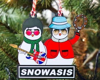 May include: A Christmas ornament featuring two cartoon penguins dressed as musicians. One penguin holds a guitar with a Union Jack design, the other a tambourine. The ornament has the text "SNOWASIS" on a black and white sign.