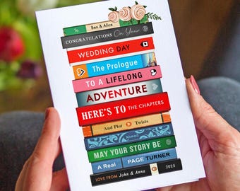 May include: A wedding card with a stack of colorful book spines. The card reads "Congratulations On Your Wedding Day" and "To a Lifelong Adventure." The card is personalized with names and a date, and decorated with floral accents.