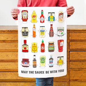 Peut inclure: Serviette de cuisine blanche avec une illustration humoristique de divers flacons et pots de condiments avec des visages. Le texte "MAY THE SAUCE BE WITH YOU" est imprim&eacute; en bas.