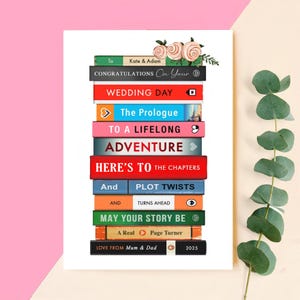 Może przedstawiać: Stos książek z tytułami takimi jak "Wedding Day" i "Adventure", ułożonych w kształt wieży. Na g&oacute;rnej książce widnieje napis "Congratulations On Your" i zdobienia kwiatowe. Na dolnej książce widnieje napis "Love From Mum & Dad 2025".