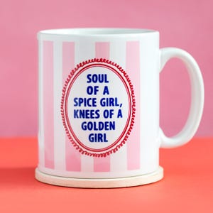 May include: White ceramic mug with pink vertical stripes and a red oval design. The text inside the oval reads "SOUL OF A SPICE GIRL, KNEES OF A GOLDEN GIRL." The mug sits on a light-coloured coaster.