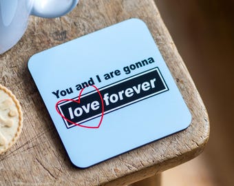 Pode incluir: Um porta-copos quadrado azul-claro com o texto impresso em preto "You and I are gonna love forever", onde a palavra "love" está dentro de um retângulo preto e um contorno fino de coração vermelho desenhado à mão rodeia a palavra "love".
