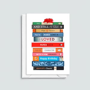 May include: A greeting card featuring a stack of colorful books with titles like "AND STILL A Total" and "Happy Birthday." A red bow sits atop the stack. The card is white with a white envelope.