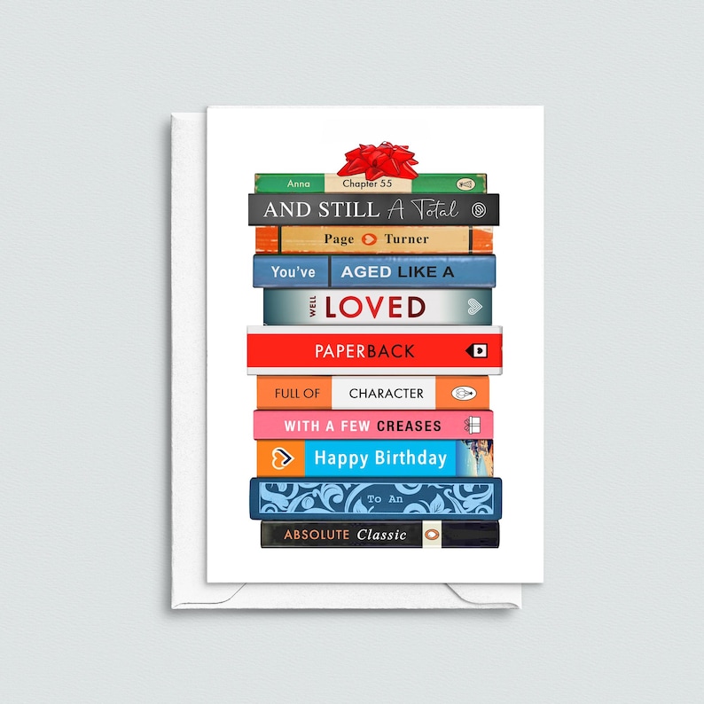May include: A greeting card featuring a stack of colorful books with titles like "AND STILL A Total" and "Happy Birthday." A red bow sits atop the stack. The card is white with a white envelope.