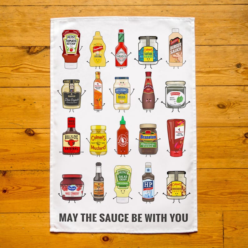 Peut inclure: Serviette de cuisine blanche avec une illustration color&eacute;e de divers flacons et pots de condiments avec des visages de dessins anim&eacute;s. Le texte "MAY THE SAUCE BE WITH YOU" est imprim&eacute; en bas.