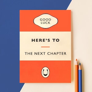May include: A greeting card with a retro design. The card is orange and white with the words "GOOD LUCK" in a white oval, and "HERE'S TO THE NEXT CHAPTER" in black. A horseshoe graphic is at the bottom. Coloured pencils are in the background.