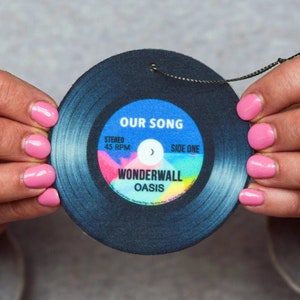 May include: A black vinyl record car air freshener with a colourful design. The record features the text "Our Song", "Wonderwall", "Oasis", "Stereo", "45 RPM", and "Side One".